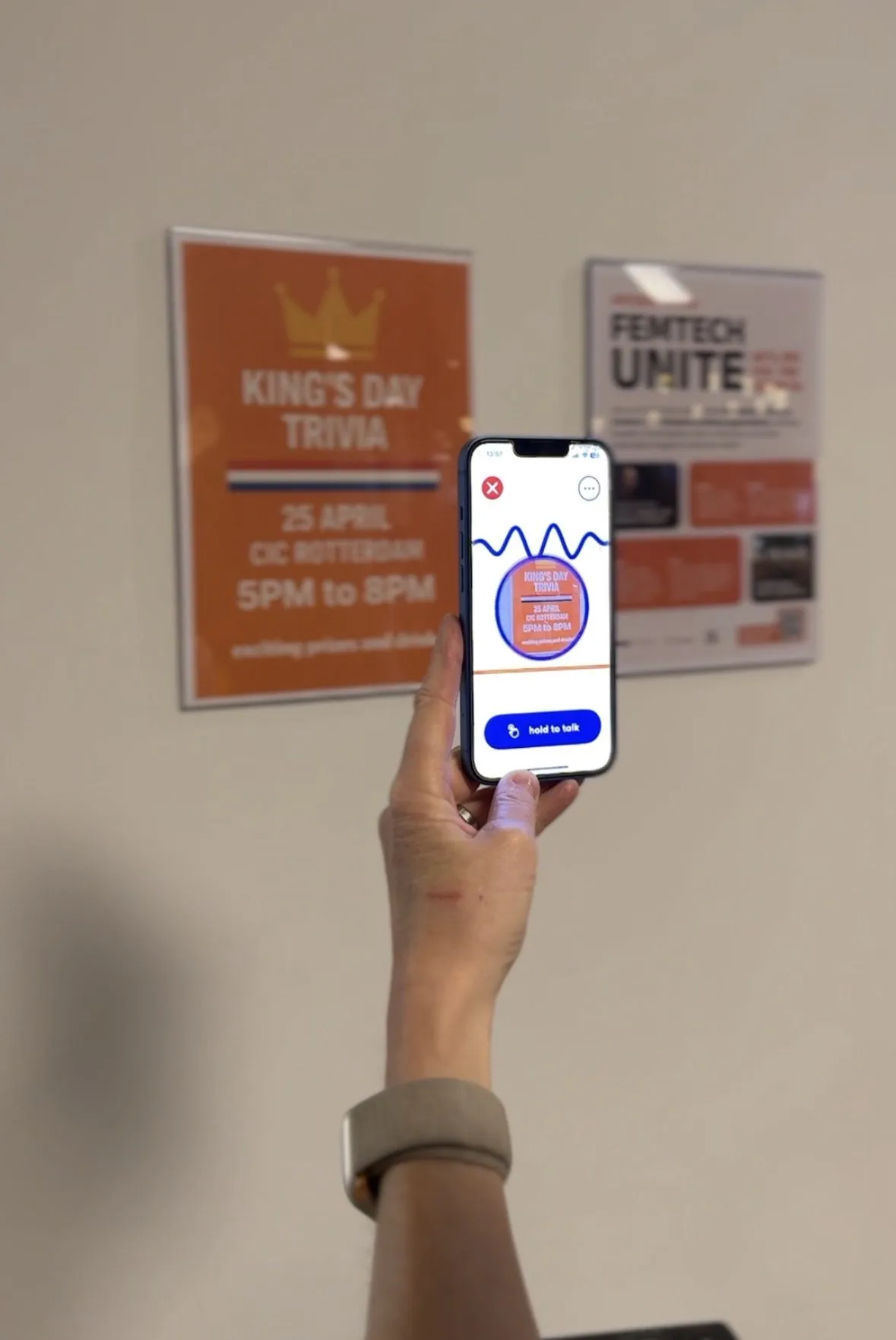 A hand holding a phone up to two event posters on an office wall, Ally showing what it can see and waiting on a hold to talk button.