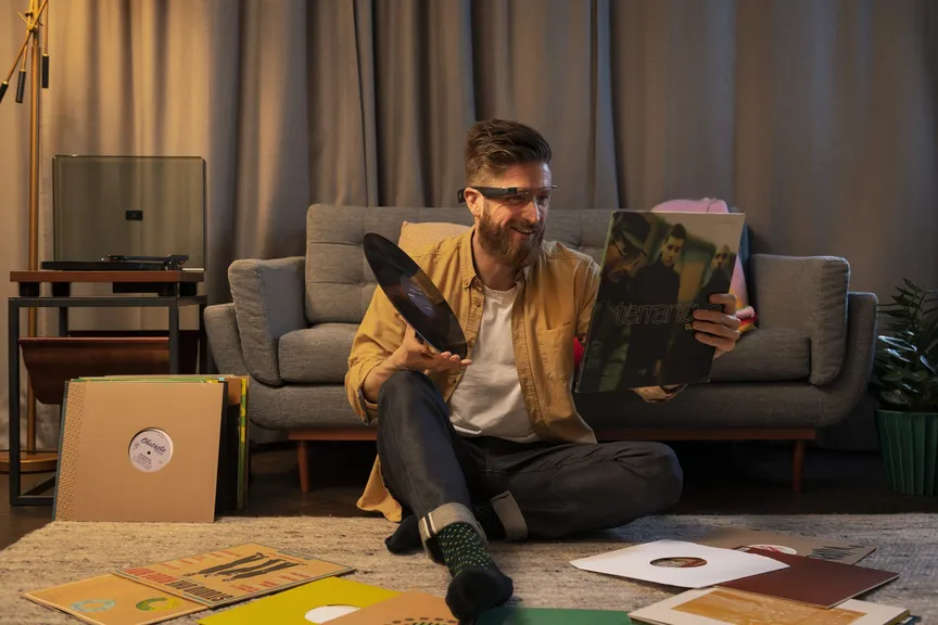 A man sitting cross-legged on his living room floor among sleeves of vinyl, holding a record up in one hand and its cover in the other while the glasses read it to him.