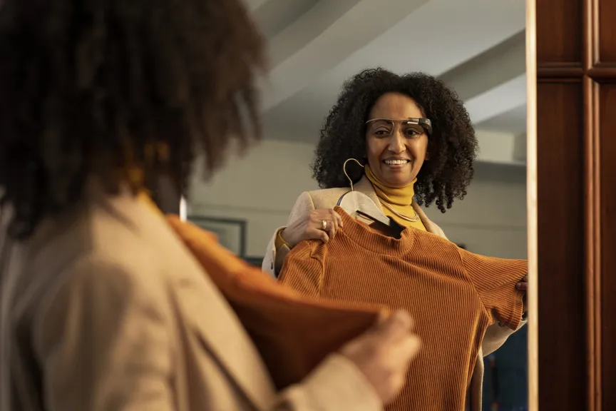 A woman holding up a rust-coloured knitted top on a hanger to show a friend, deciding on it with the glasses on.