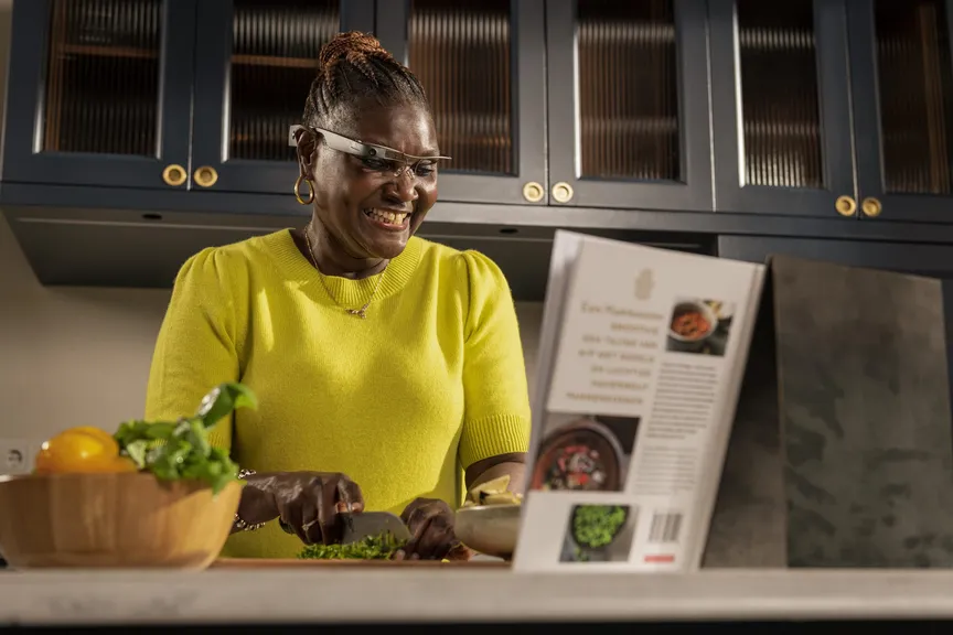A woman in a bright yellow jumper chopping herbs at a kitchen counter, grinning, an open cookbook propped up beside her.