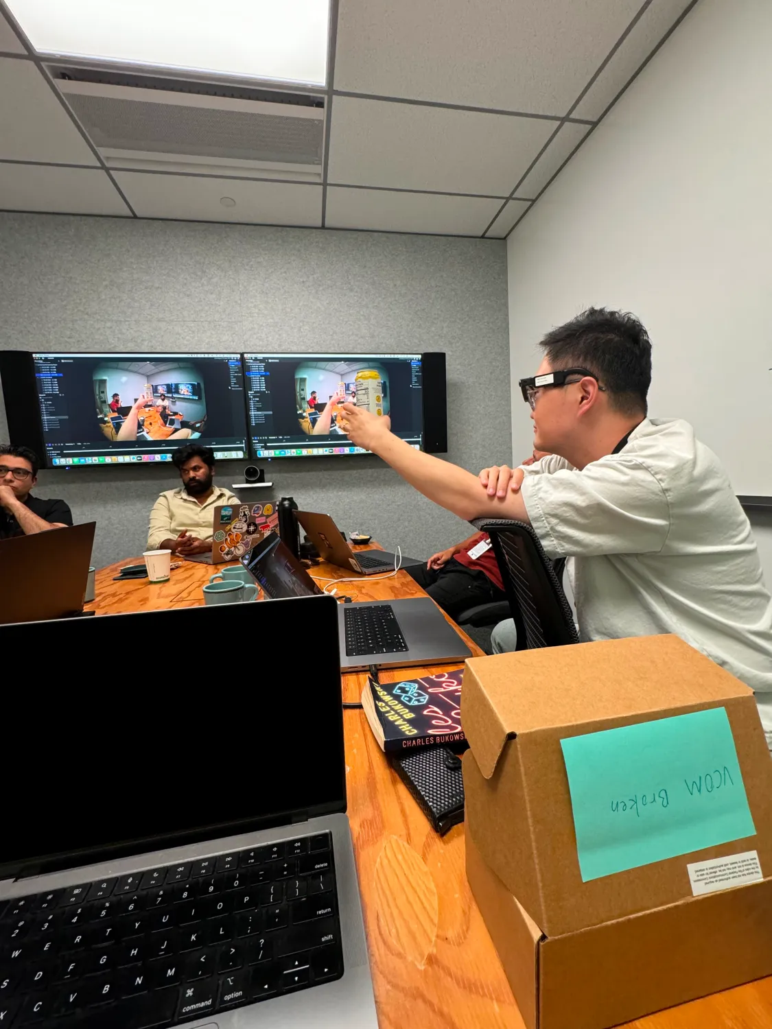 A group of four people are gathered in a modern conference room around a wooden table with laptops. One person on the right, wearing AR glasses, gestures toward a large screen on the wall that shows a mirrored camera feed of the meeting.