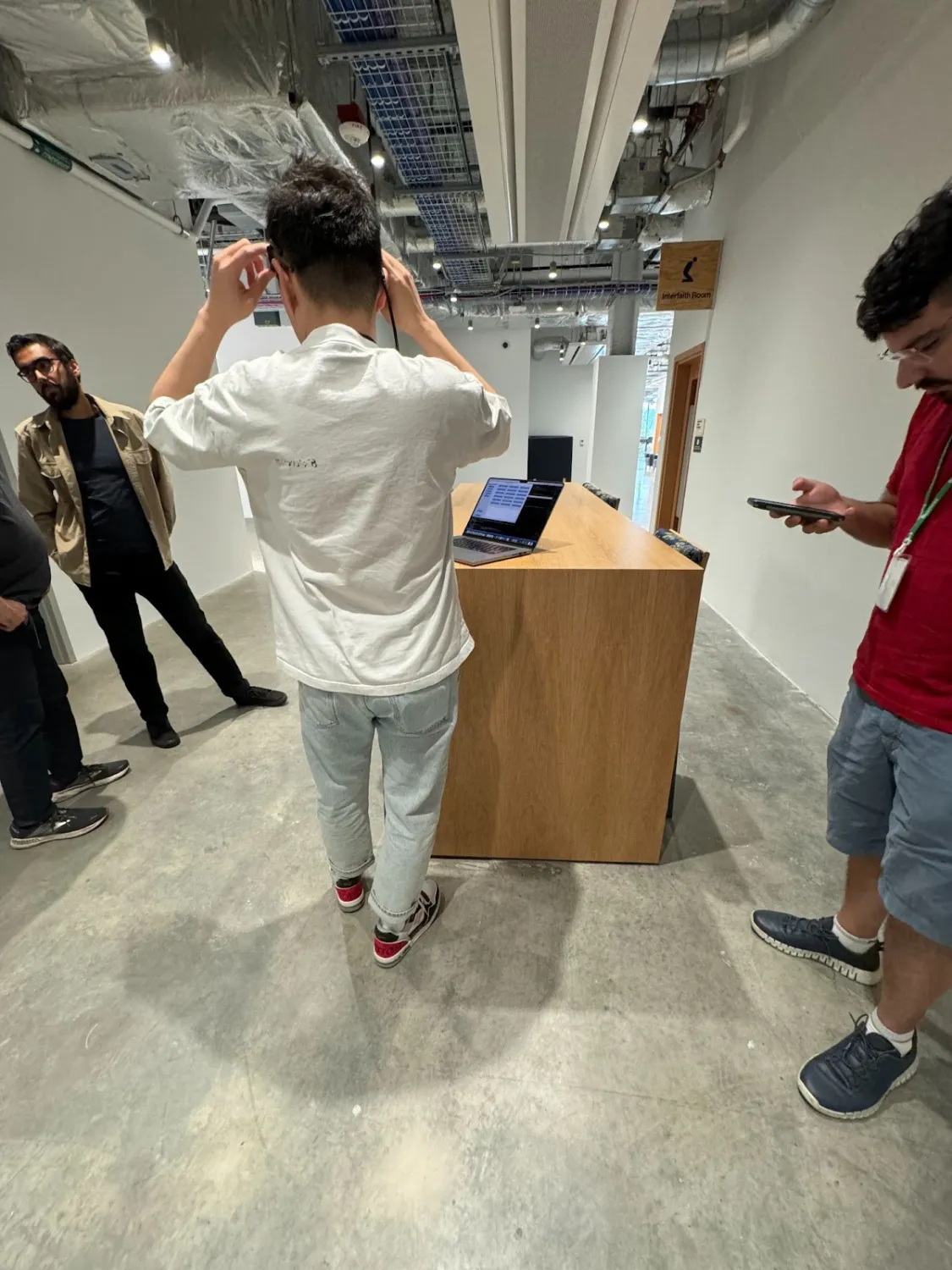 A person in a white shirt and light jeans is seen from behind adjusting a pair of smart glasses in a modern industrial-style hallway with exposed ducts and lighting. Three other people stand nearby — one looking at a phone, two engaged in conversation. A wooden kiosk-style desk holds a laptop displaying a dark interface.