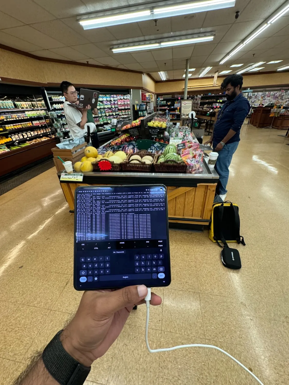 A person holds a foldable tablet displaying a terminal interface with lines of code or logs. In the background, one man stands with a laptop while another observes the fruit display, which includes melons, apples, and packaged produce.