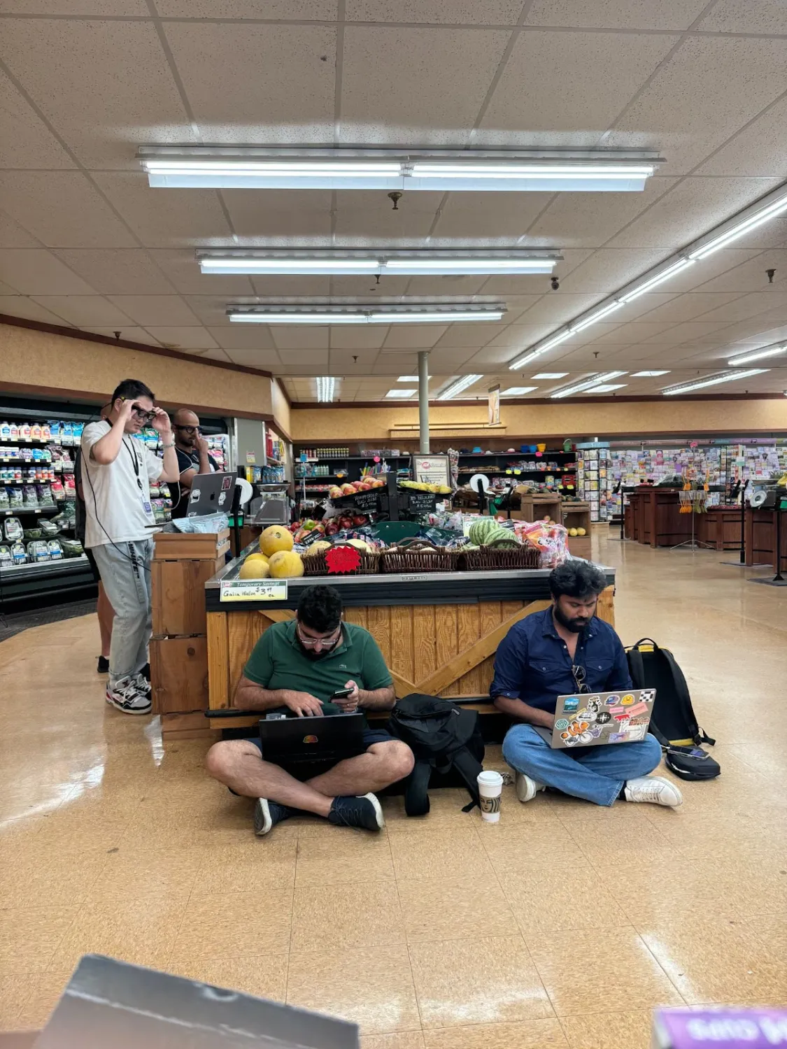 In a supermarket produce section, two men sit on the floor working on laptops in front of a display of fruits, including watermelons and melons. Behind them, another man adjusts a pair of smart glasses while a colleague watches nearby with a laptop on a wooden stand. Coffee cups and backpacks are scattered around, indicating a casual yet focused tech setup in an unconventional setting.