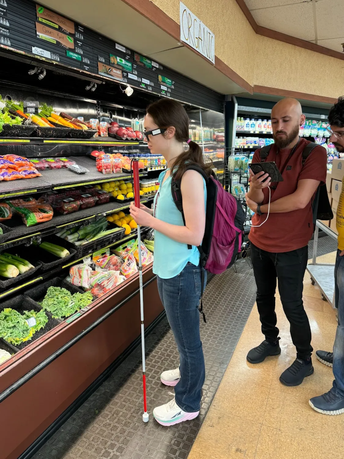 A woman wearing smart glasses and holding a white cane stands in front of a vegetable display. Two men stand nearby, one monitoring something on a smartphone connected by a cable supporting the smart glasses setup. The produce shelves behind them are stocked with fresh vegetables like carrots, kale, lettuce, and cucumbers.