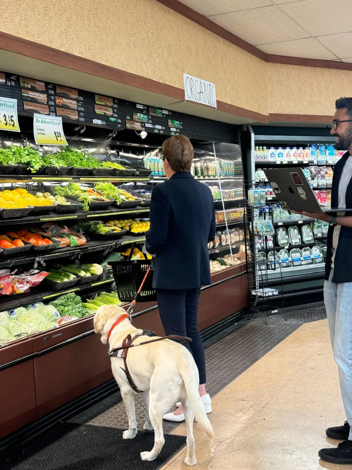 A woman with a guide dog stands in front of a produce display in the organic section of a supermarket, facing shelves stocked with lettuce, cilantro, peppers, and other vegetables. She is accompanied by a man nearby holding an open laptop. The guide dog, a yellow Labrador, is wearing a harness and looks attentively ahead.