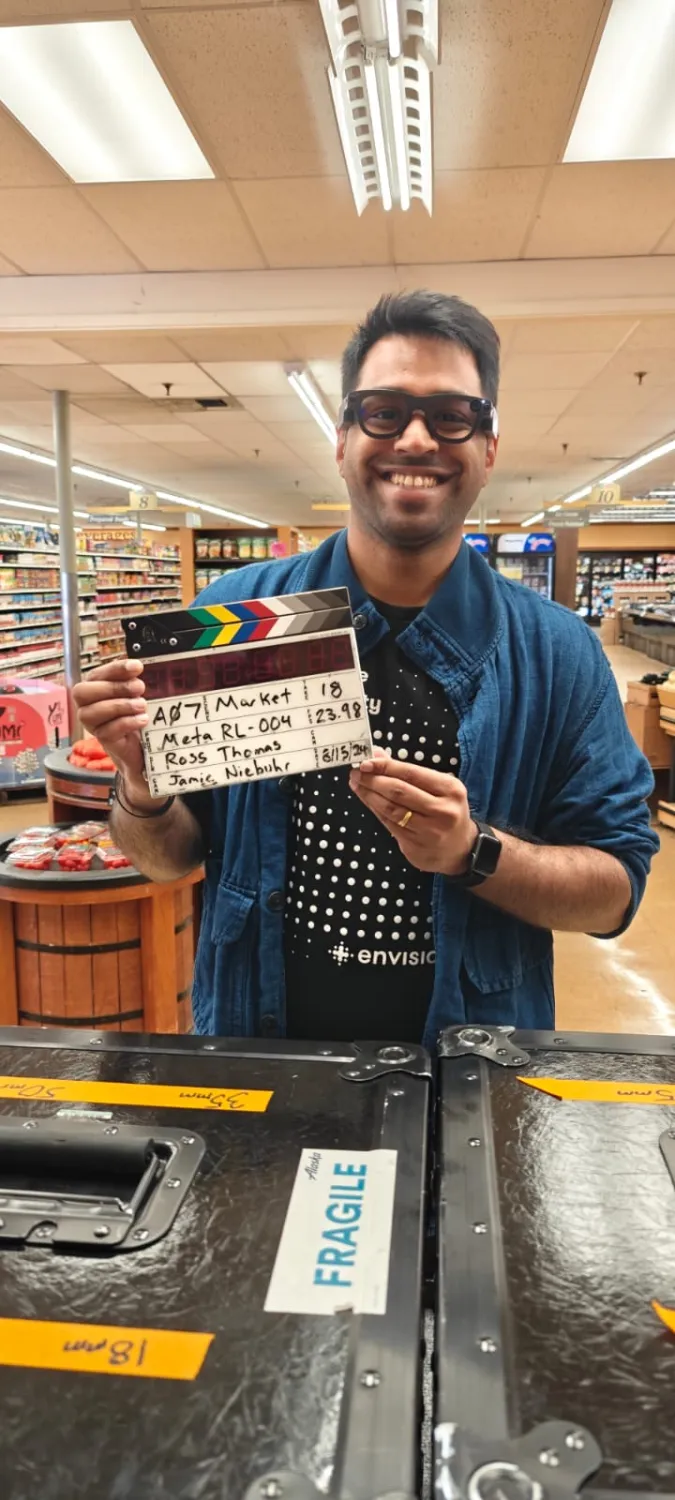 A picture of me wearing the Aria Gen 2 glasses inside a supermarket holding a film slate labeled “Meta RL-004” with details including the names and dates of filming. I’m wearing a dark dotted shirt with the Envision logo and a blue jacket. In front of me are two black equipment cases marked “FRAGILE” and labeled with orange tapes.