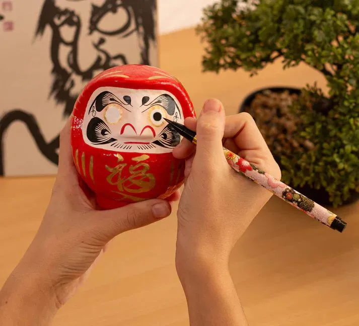 A hand painting the first eye onto a red daruma doll with a brush, the wish being made