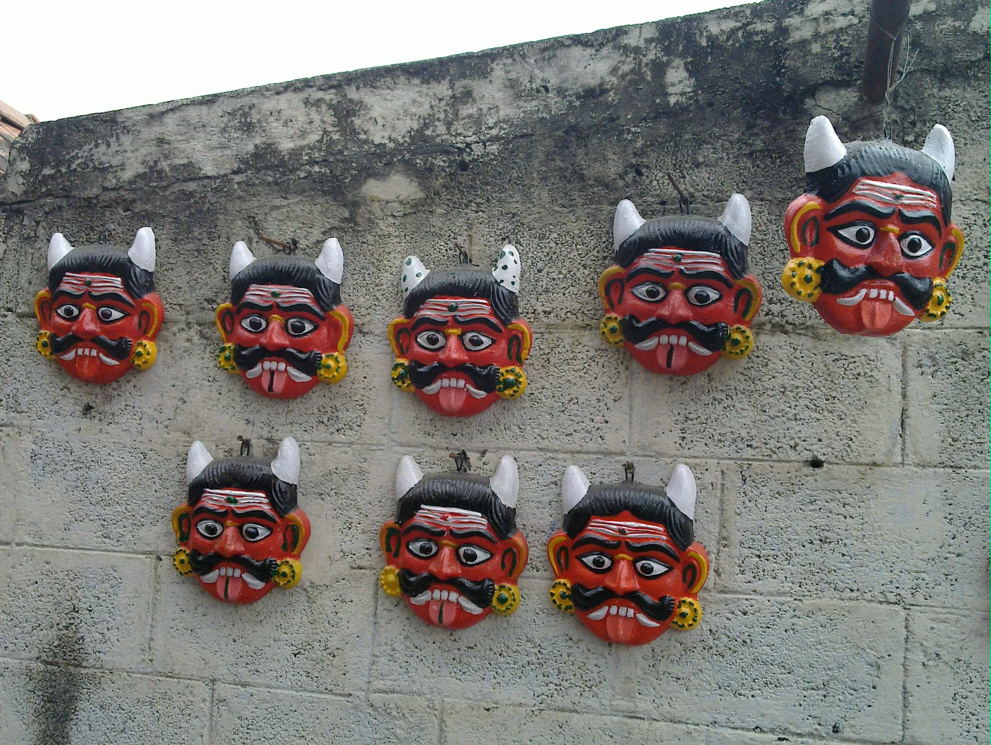 Painted drishti bommai masks hanging in rows on a whitewashed street wall, red faces with horns, moustaches and tongues out
