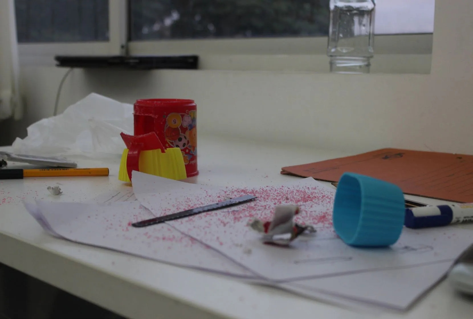 The studio desk mid-prototype: a red plastic cup with its handle sawn off, a yellow silicone band, pink plastic shavings scattered across the paper and an empty jar on the windowsill behind