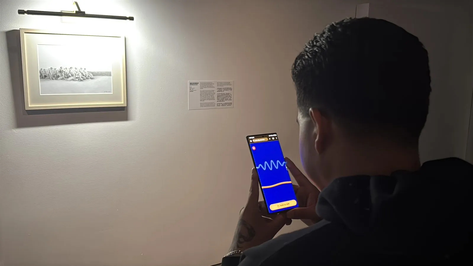 A visitor in a dimly lit gallery holding his phone towards a framed black and white photograph, the Ally waveform bright on the screen.