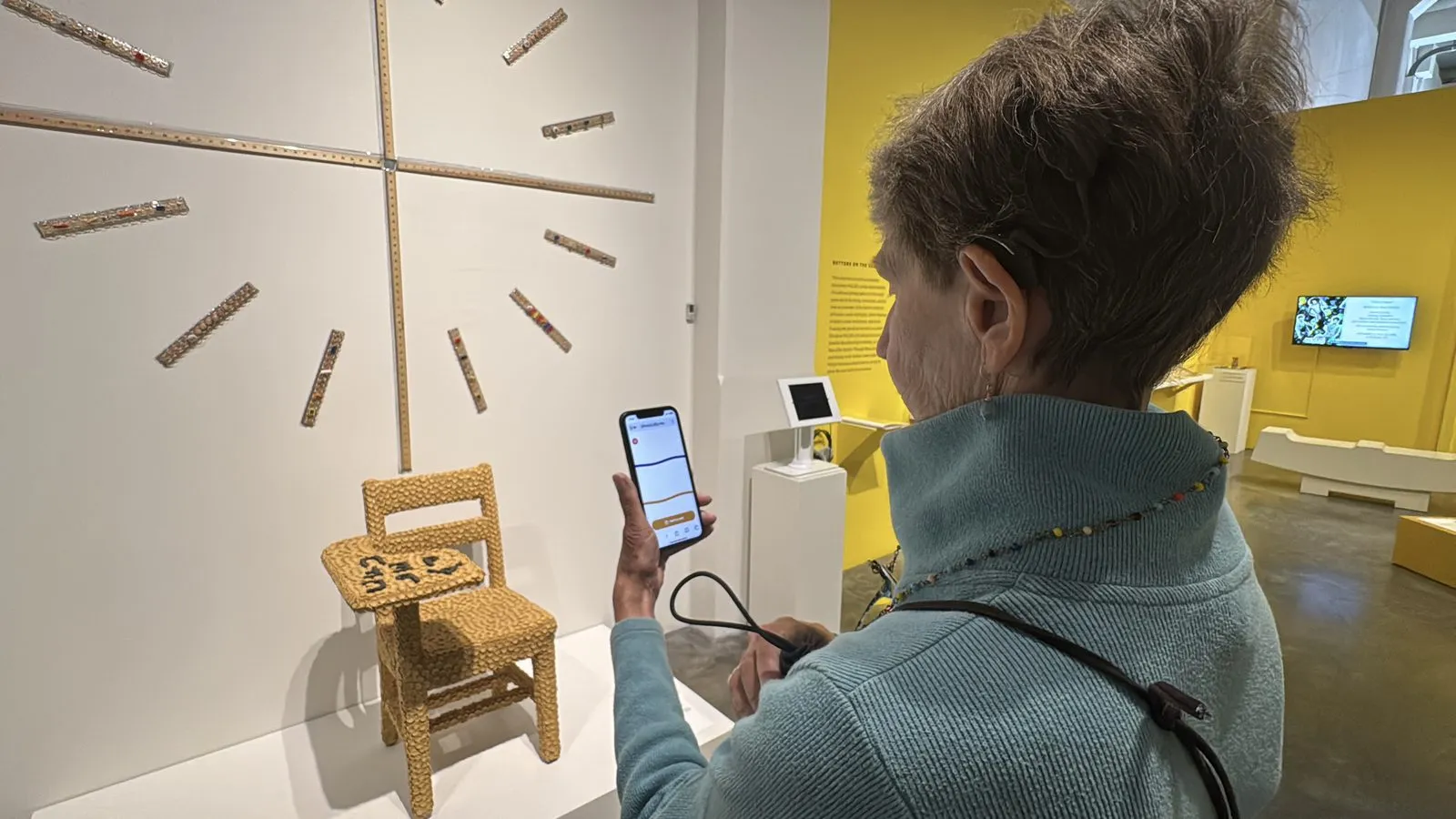 A visitor at the Museum of Craft and Design holding her phone up to a crocheted chair on a plinth, Ally listening on screen, a yellow gallery wall behind her.