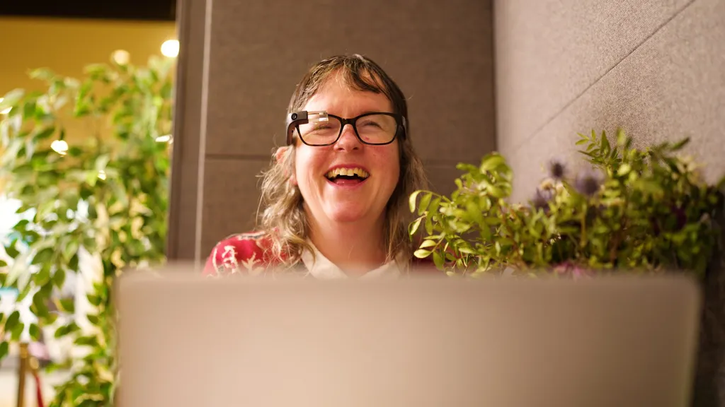 A woman laughing at her laptop screen, wearing Envision Glasses over her own spectacles, on a call with a sighted companion.