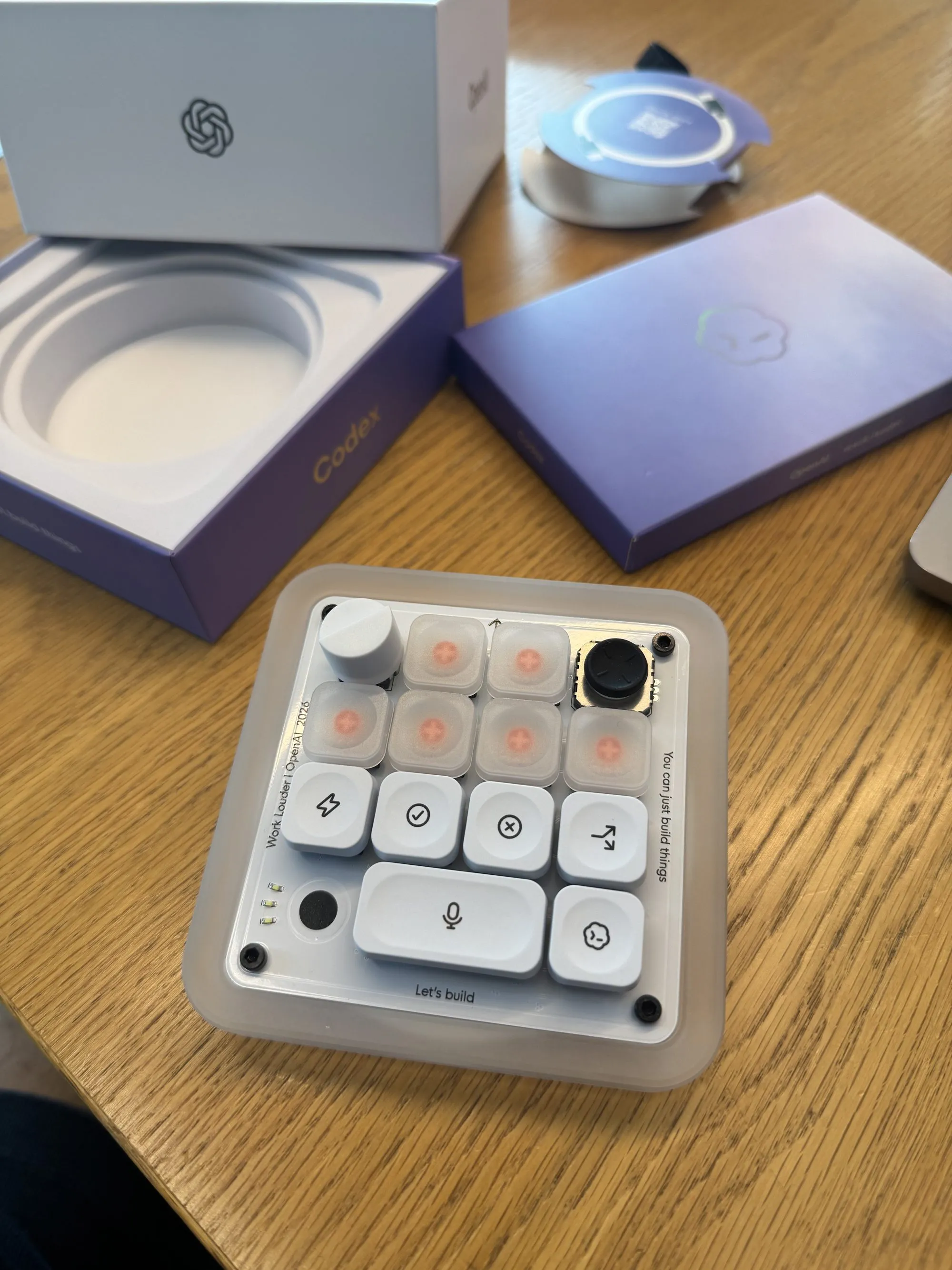 The keypad out of its box on my desk: a compact translucent-cased grid of white keys, a knob and a joystick along the top row, a wide microphone key across the bottom, and accept, reject and lightning keys in between. “Work Louder | OpenAI 2026” is engraved down one edge and “You can just build things” down the other, with the purple Codex packaging still open behind it.