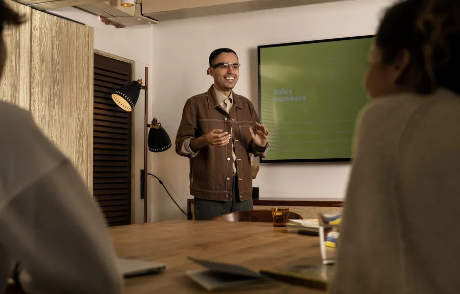A man standing at the head of a meeting room table presenting a slide of sales numbers, colleagues in the foreground, glasses on.