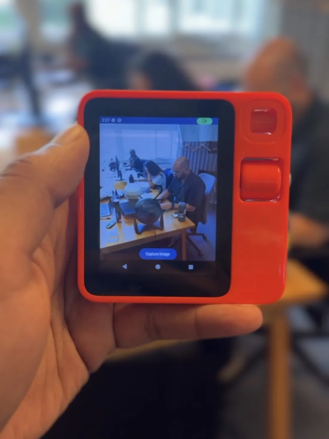 The Rabbit R1 held in one hand, its little screen showing a captured photo of the room.