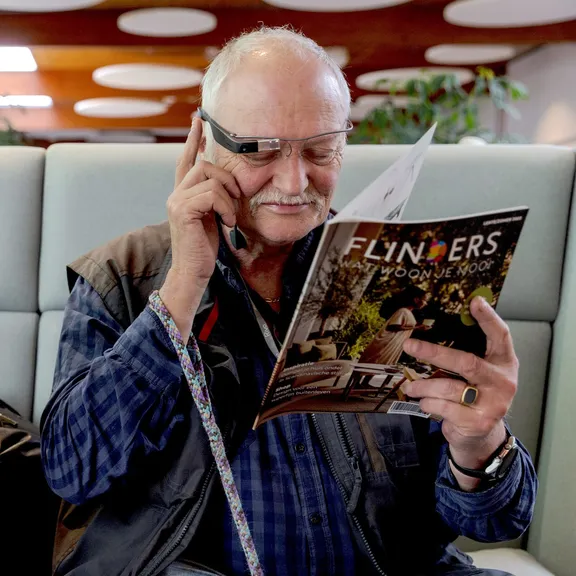 An older man sitting on a pale green sofa with an interiors magazine open in both hands, one finger resting on the temple of his Envision Glasses while it reads the page to him.