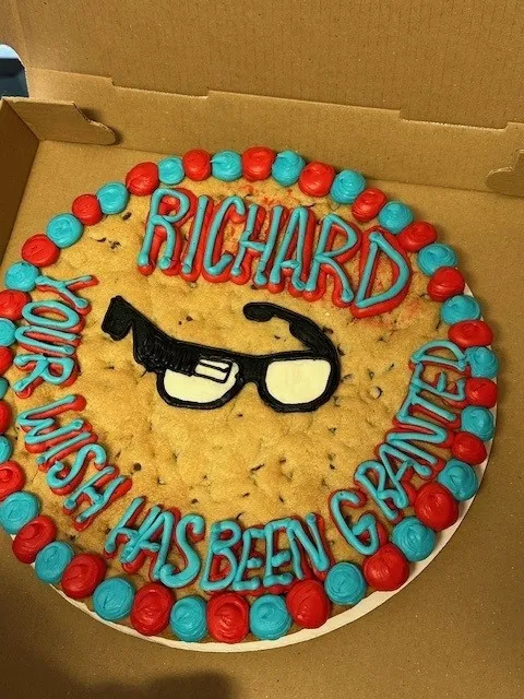 A large cookie cake with colorful red and blue icing that reads “RICHARD” at the top and “YOUR WISH HAS BEEN GRANTED” around the edges. The center of the cake features a black and white design of glasses. The cake is in an open cardboard box.