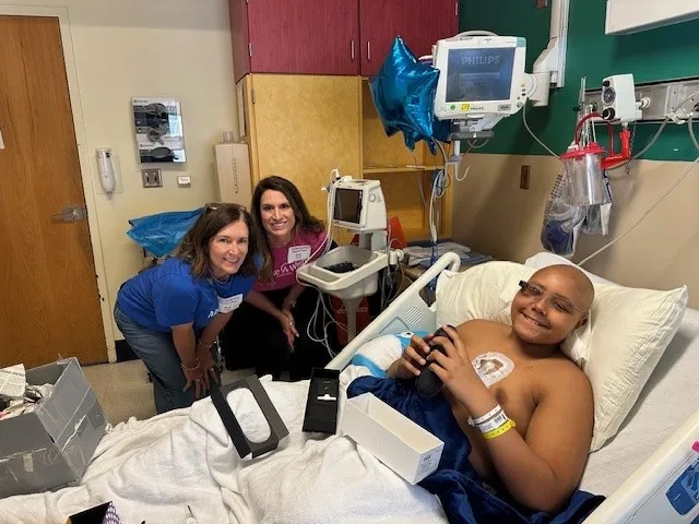 A young boy with a shaved head lies in a hospital bed, smiling, while holding assistive glasses in his hand. Two women, both smiling, stand beside him. The room has medical equipment, blue balloons, and boxes, indicating a celebratory moment.