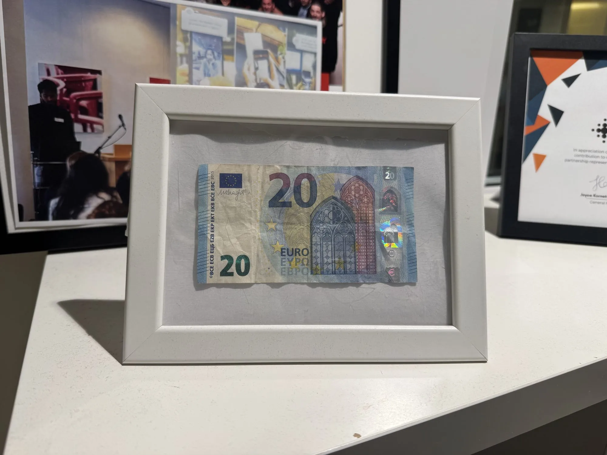 The twenty euro note itself, creased from her purse, in a plain white frame on a shelf in our office next to team photos and a partnership award.