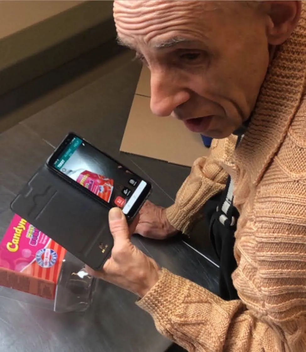 An older user in a shop, leaning in over an Android phone held above a bright pink box of sweets, listening to the app read the label back to him.