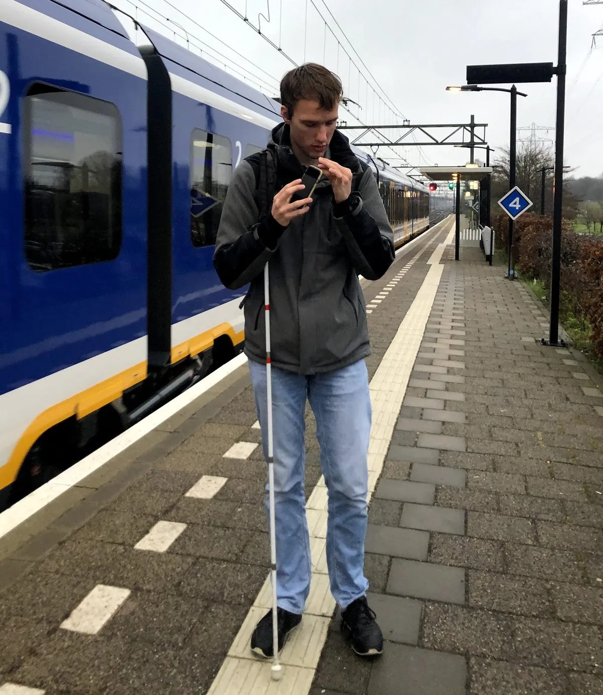 A user on a train platform in the Netherlands, white cane in one hand, holding his phone up close to hear what the app makes of the departure board while a train pulls in beside him.