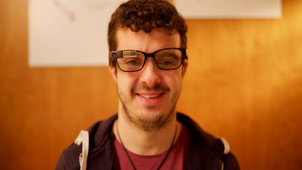 A man smiling straight at the camera, the Envision Glasses frame clipped in front of his own thick-rimmed spectacles.